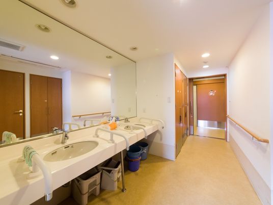 壁一面の鏡の前に洗面ボウルがいくつか設置してある。それぞれの洗面ボウルの両サイドに手すりがある。足元は空間である。