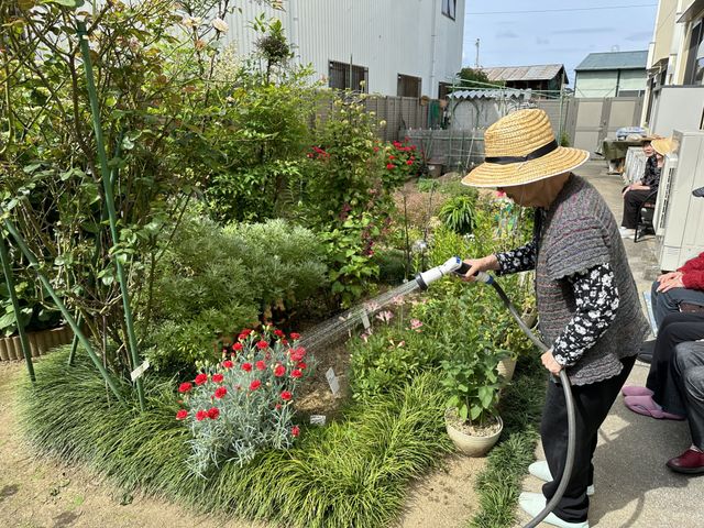庭園で植物に水やり 施設敷地 手入れが行き届いた庭で、利用者が植物の水やりをして心穏やかな時間を過ごしています。庭仕事は心身の活性化に役立つ穏やかな日課です。