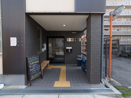 黄色い点字ブロックが、玄関ポーチの床に取り付けられている。右側には飲料の自動販売機があり、左側には木製のベンチが置いてある。