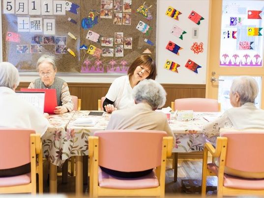 スタッフと入居者様達が席に着いている。壁には鯉のぼりの折り紙飾りや写真がたくさんあり、賑やかな空間である。