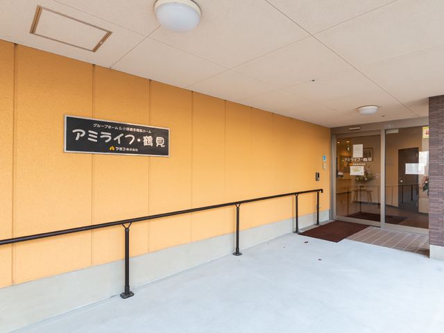 黄色の壁とエントランス看板 玄関 明るい色の壁が迎えるエントランスは、訪れる人々を温かく迎えています。ご高齢の方々が毎日の生活を送る場として、あなたの手で支えてみませんか。
