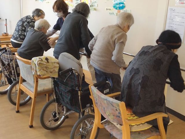 高齢者が並ぶ訓練室 機能訓練室 明るい空間で、サポートを受けながら体を動かす機能訓練の様子です。笑顔と活気に溢れています。
