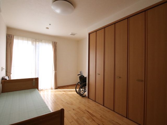 木目調の洋室と車いす 居室 明るく広々とした洋室が見受けられ、快適な個室空間が確保されています。車いすも置かれており、バリアフリーへの配慮が行き届いている様子です。