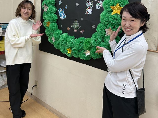 掲示物前のスタッフ 掲示物   季節感のある掲示物の前で、スタッフの方が笑顔で案内しています。手づくり感のある装飾があり、温かな雰囲気が伝わります。