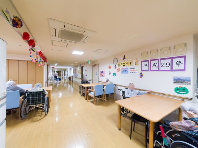 廊下の様子 談話コーナー兼食堂  広く明るい廊下には、車椅子の方々がゆったりと過ごしており、壁には季節の装飾が施されています。お一人お一人の安全と快適な時間を大切にした環境が整っております。