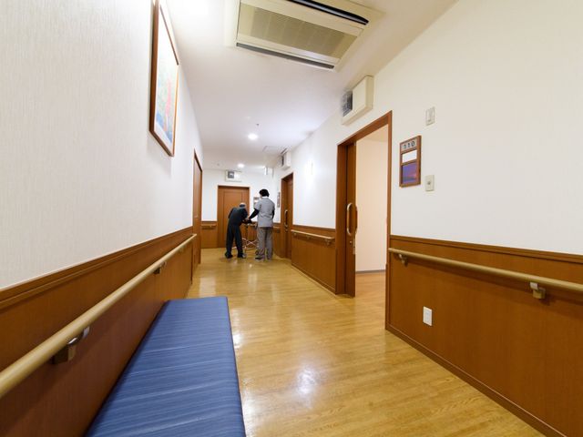 廊下でスタッフと利用者 通路 落ち着いた雰囲気の廊下で、スタッフが利用者様の歩行支援をしています。ゆったりとした空間で安心してお過ごしいただけます。