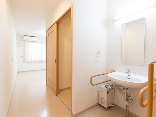 施設の写真 白い居室に合ったシンプルな洗面台。幅は広く取られており両側にしっかりとした手すりもついている。大きい鏡なので見やすくなっている。