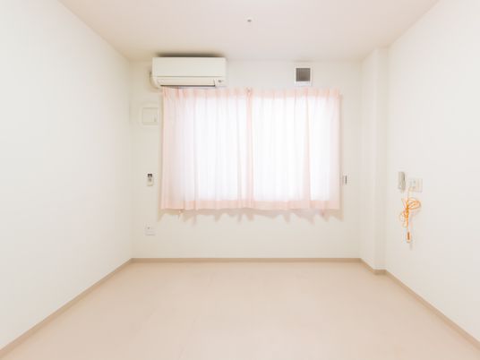 施設の写真 白色で揃えた清潔感のある居室。エアコンがある。長いコード付きの緊急用ボタンが壁に備え付けてある。