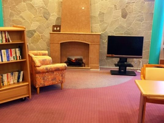 正面の壁は石造りで暖炉を模した暖房器具がある。横にはテレビがあり、一人掛けのソファが２脚並んでいる。ソファの後ろには本棚がある。