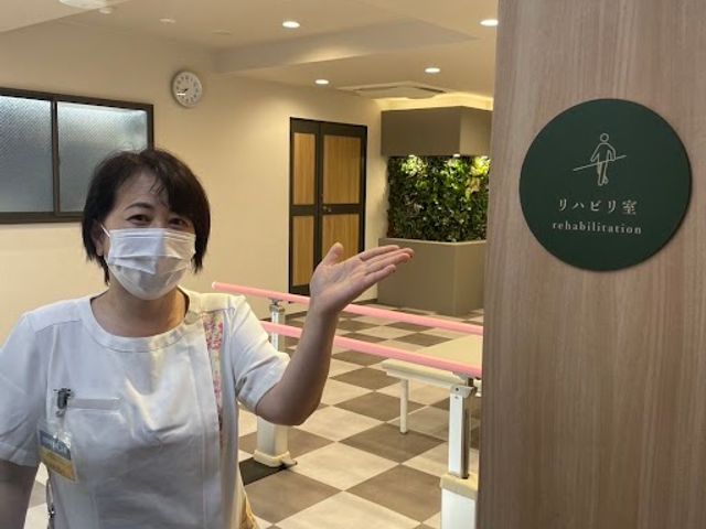 リハビリ室の案内 機能訓練室   明るい空間に平行棒が設置されたリハビリ室です。スタッフの方が案内しており、利用者の支援しやすい雰囲気が伝わります。