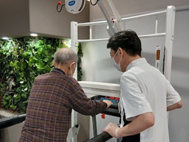 機能訓練を受ける様子 機能訓練室   明るい機能訓練室で、スタッフの方が利用者の方を支えながら器具の前へ案内しています。安全に運動しやすい環境づくりが伝わります。