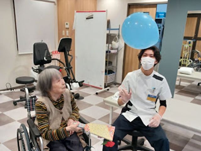 機能訓練室 機能訓練室   車椅子の方とスタッフが見え、明るい訓練スペースで会話しながら過ごしています。運動器具もあり、日々の支援の様子が伝わります。