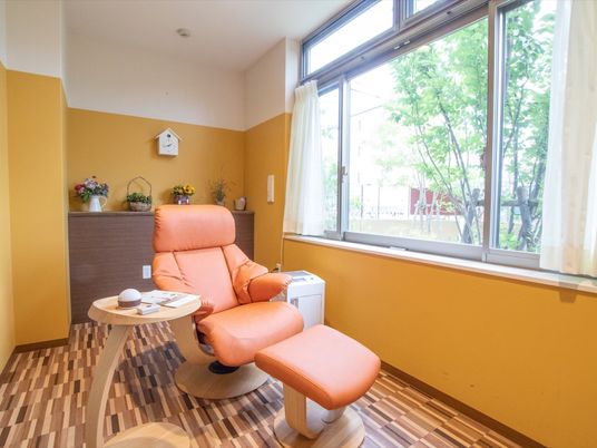 大きな窓が開いた個室に、オレンジ色のリクライニング付きソファと足置きが置かれている。壁の棚に花が飾られている。