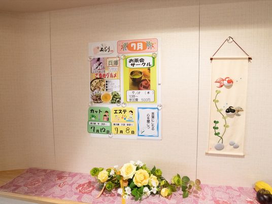 施設で開催されるレクリエーションやエステ、カット、ご当地グルメの広告などが壁に貼りだされている。棚に花が飾られている。