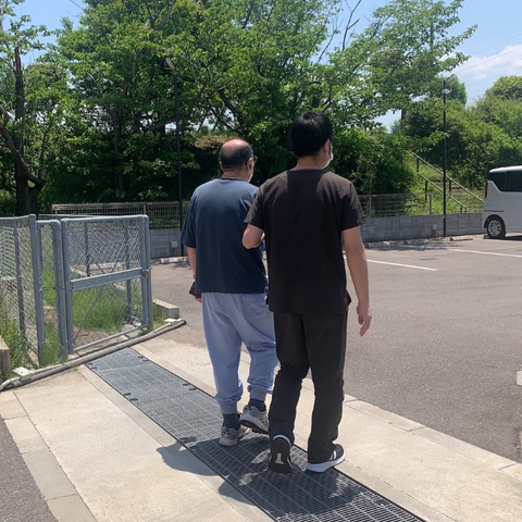 車椅子の利用者とスタッフの歩行 外観 スタッフが利用者の方と一緒に歩いている様子です。屋外で緑豊かな風景が広がります。