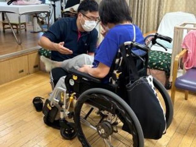 車椅子の介助 機能訓練室 車椅子の利用者に向けた親身なサポートを行っています。木目の床が温かみを感じさせます。
