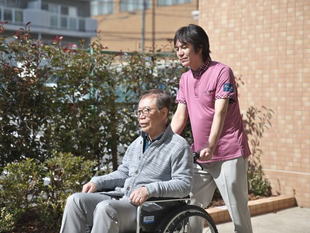 車椅子を押すスタッフと利用者の様子 庭 スタッフが利用者の車椅子を押し、和やかに屋外を散歩中。リラックスした時間が流れます。