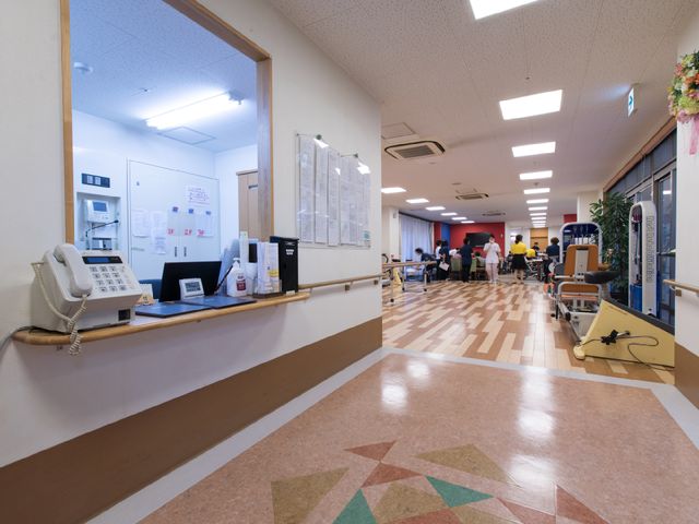 広々とした廊下の景色 機能訓練室近くのスタッフルーム ゆったりとした廊下には、利用者が快適に過ごせる座席や訓練器具が見られ、スタッフや利用者の皆様が穏やかに交流されています。