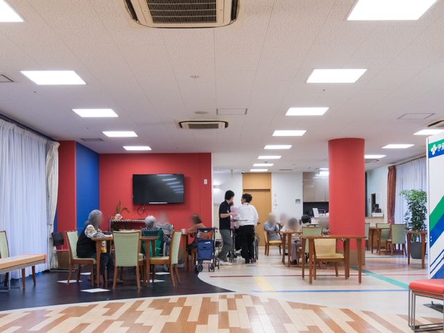 広々とした明るい食堂 機能訓練室から見た食堂 ゆったりとした椅子に座る利用者様が食事を楽しんでおり、スタッフが丁寧にサポートをしています。交流が育まれるアットホームな雰囲気が感じられます。