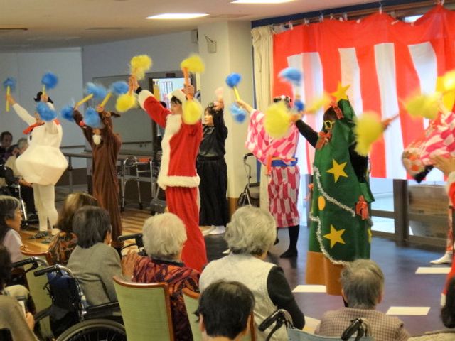 イベント風景の活気ある共有スペース クリスマス カラフルな衣装に身を包んだスタッフがイベントを行い、車椅子に座る方々が楽しそうに見守る和やかな空間です。
