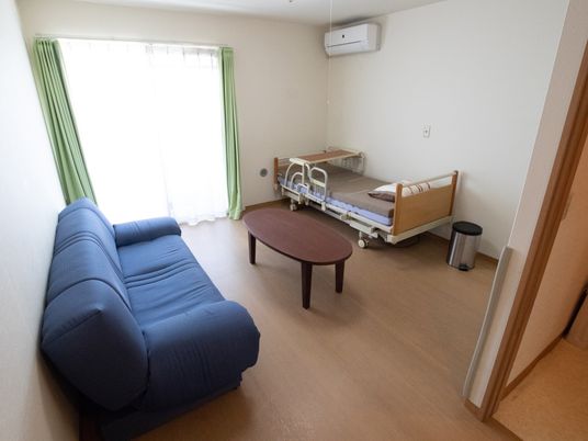 奥にはエアコンと足元に机のあるベッドとごみ箱が設置してある。部屋の中心に机があり、手前側に青いソファがある。