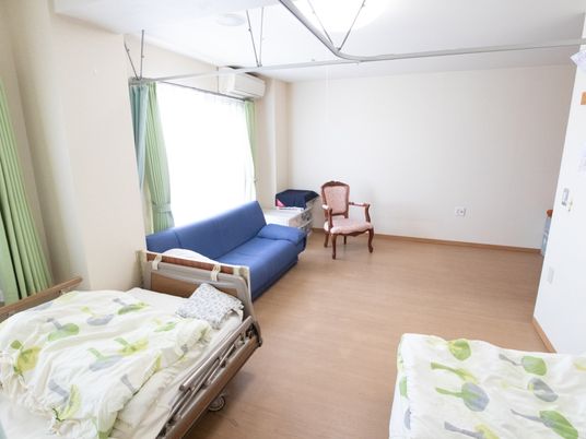 施設の写真 手前には二人分のベッドがあり、左に仕切るためのカーテンがある。また、青色の大きいソファと収納ケース、椅子がある。