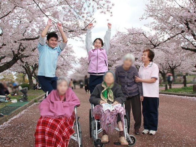 桜並木を背景にした施設利用者との交流 庭 桜の下で、利用者とスタッフが和やかに身体を動かしています。季節を感じられる場所です。