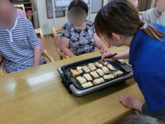 軽食を作る様子の居室 居室 テーブルで軽食を準備中です。利用者とスタッフが、和やかに交流しながら料理を楽しむ場面です。