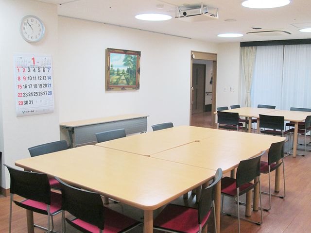 明るい食堂の様子 地域交流室 ゆったりとした食堂にはテーブルと椅子が配置され、壁には暦と絵画が掲げられています。