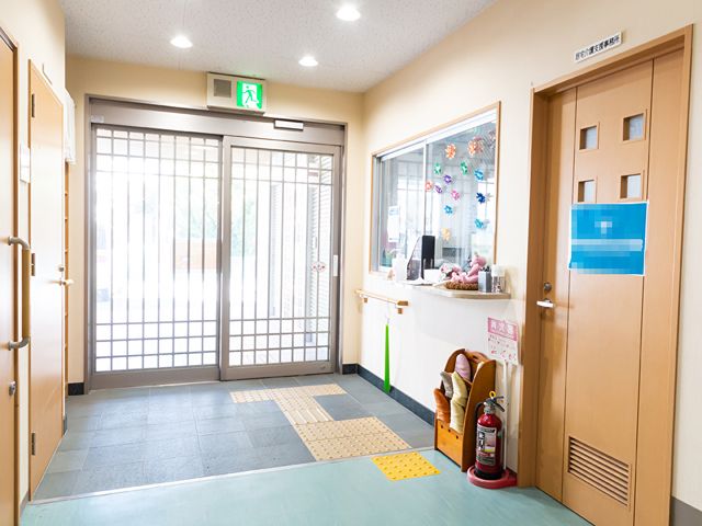 明るいエントランスホール 玄関 広々とした入り口には車椅子用のスペースもしっかりと設けられ、安全を考慮した設計が見受けられます。フロントカウンターには優しい雰囲気の手作りの飾りがあり、訪れる人を暖かく迎えてくれそうです。