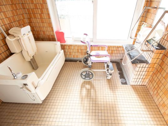 大きい窓がある。浴室用の車椅子が置いてある。洗い場に手すりが設置してある。介護浴槽になっている。床と壁がタイルで敷き詰められている。