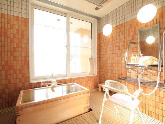 窓から光の差す明るい浴室である。浴槽は木で造られており、温かみを感じさせる。介護用の風呂椅子が設置されている。