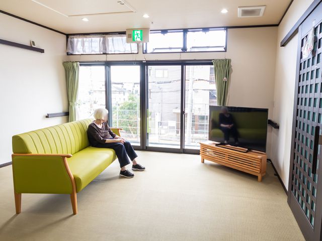 居室でくつろぐ一人 共有スペース ゆったりとしたソファが置かれたリビングで、一人が静かにテレビを観ています。明るく開放感のある空間で、リラックスした時間が過ごせそうです。
