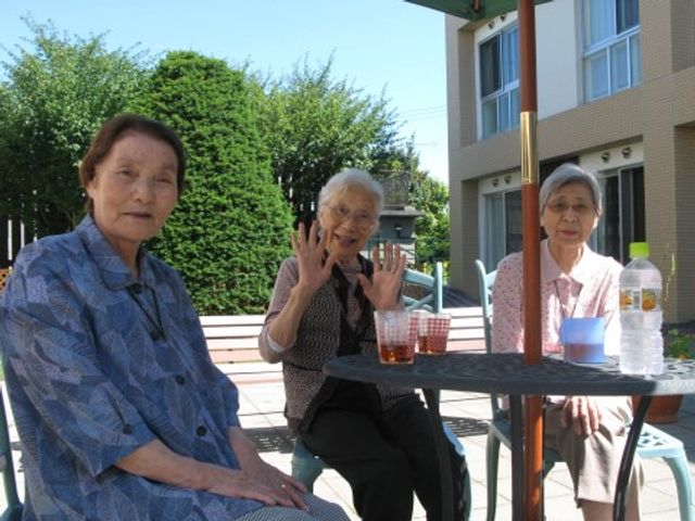 笑顔の利用者三名中庭 中庭 ゆったりとした陽気の中、中庭で椅子に腰掛けてくつろぐ利用者の様子が感じられます。お茶を楽しむ笑顔は、ここでの穏やかな時間を象徴しています。
