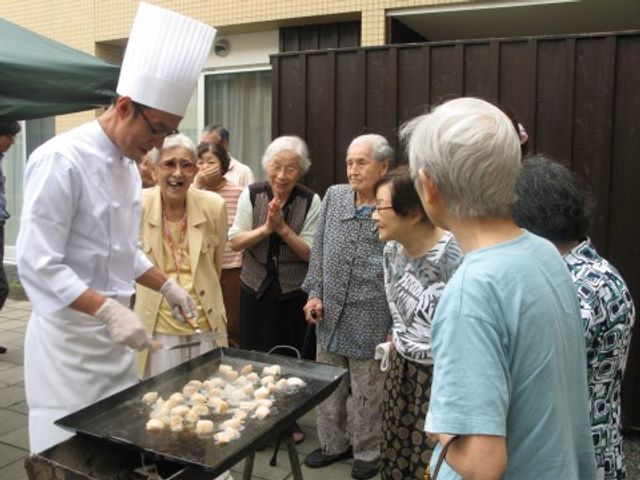 料理するシェフと笑顔の高齢者 イベント シェフが焼きたてのお団子を提供し、笑顔で囲む高齢者たち。屋外での料理イベントが彩り豊かな日常を提供しています。