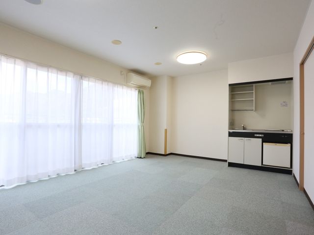 明るい居室の一角 居室 広々とした空間の居室で、大きな窓から自然光が差し込み、明るく開放的です。キッチン付きで日々の生活が快適に過ごせるよう配慮されています。
