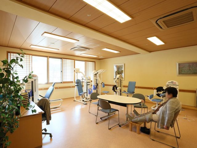 機能訓練室の日差し入る風景 機能回復訓練室 温かな光が差し込む機能訓練室で、スタッフがケアを提供しています。最新の設備が整い、リハビリに取り組む方々をサポートします。