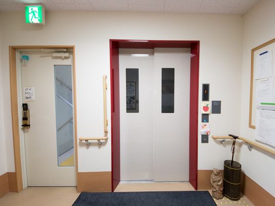 白い扉に赤いフレームのカラーが目立つエレベーターがある。車椅子専用ボタンもある。扉横には手すりがあり、つかまって待つことができる。