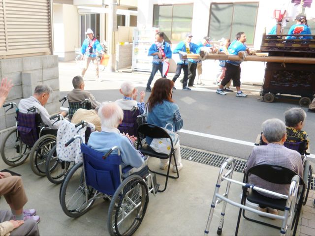 車椅子の人々とパレード イベント エントランスでは、屋外でのイベントを楽しむ利用者の方々が、笑顔で手を振っています。