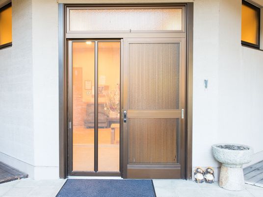 茶色い金属製の格子がはまっている玄関ドアがある。引き戸になっており、網戸が設置されている。足元には玄関マットが敷かれている。