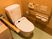 茶色い壁に囲まれたトイレがある。便器には蓋が付いており、可動式のひじ掛けが付いている。壁にはＬ字型の手すりがある。