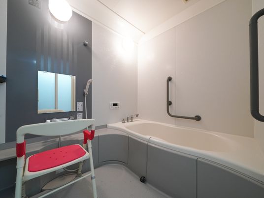 洗い場の鏡の前に、背もたれのある椅子が一脚置かれている。白い壁には黒い手すりが２箇所設置されている。