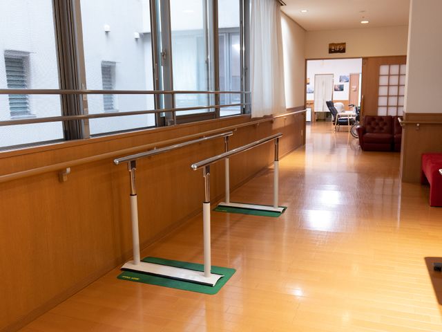 廊下の手すりと椅子 リハビリ器具A 広々とした廊下には安全に配慮した手すりが設置されており、ゆったりとした椅子も見受けられます。あたたかみのある木の色合いが落ち着いた雰囲気を演出しています。