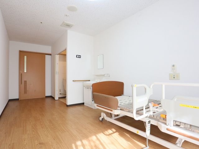 清潔な個室のベッドと入口 居室 広々として明るい個室です。利用者の自立を支援するための設備が整っており、ゆったりとした時間をお過ごしいただけます。