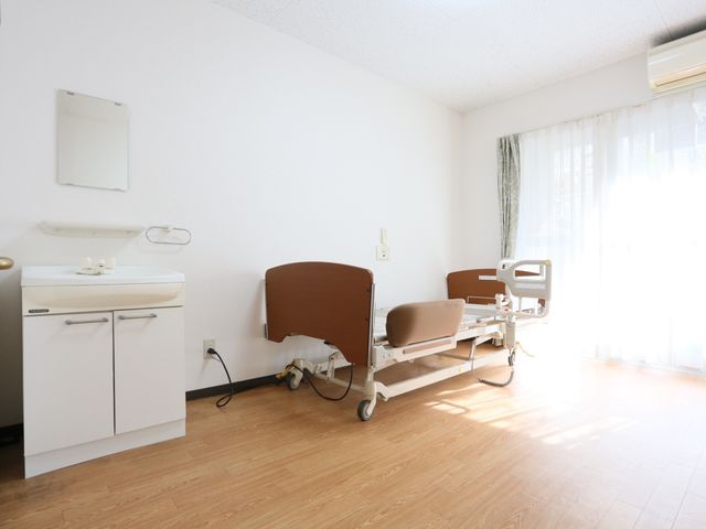 居室のベッドと洗面台 居室 床の明るい色合いが印象的な個室で、日差しを取り込む大きな窓があります。快適な生活を支える設備が整った空間です。