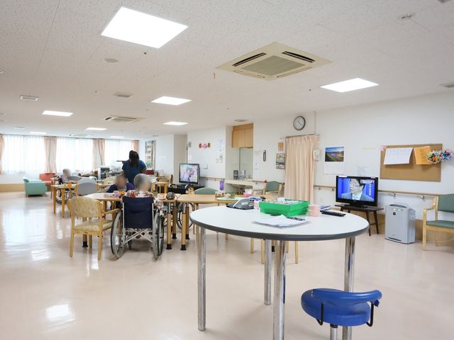 広々とした明るい居室 食堂 居室には複数の椅子とテーブルが配置され、テレビを見る方やくつろぐ姿が見受けられます。機能的な空間で働きやすさを重視しています。