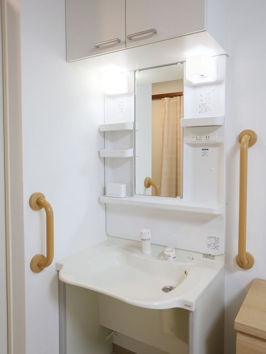 居室の白壁と同色の、清潔感のある洗面台である。たっぷり仕舞える収納棚に鏡、照明、コンセントの差込口が設けられている。