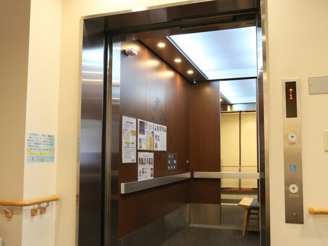 施設内の明るいエレベーター エレベーター 清潔感あるエレベーターは、ガラスと木目で落ち着いた雰囲気を醸し出しています。ゆったりとした空間設計は、利用者の快適な移動を支えます。