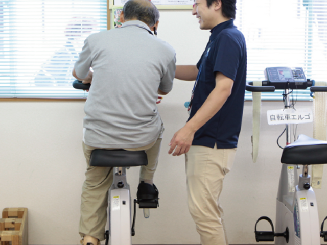 自転車型エルゴ施設 機能訓練室 スタッフがエルゴでの運動をサポートしています。利用者とのコミュニケーションが活発です。