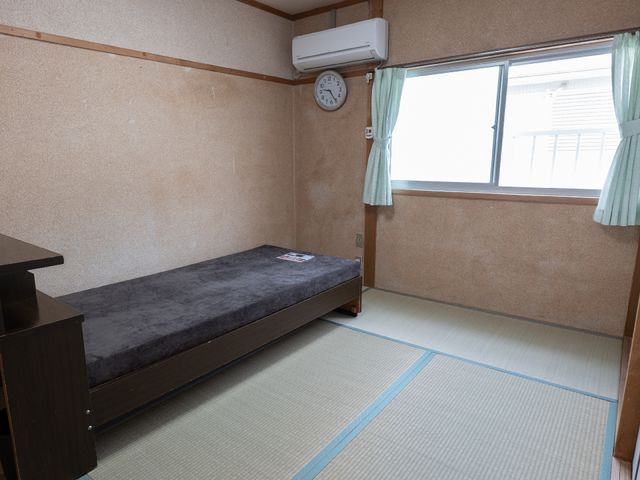 和室の居室風景 居室 清潔感のある居室は、畳の上にベッドと時計を備え、落ち着きある空間を提供しています。窓からは自然光が差し込み、穏やかな時間を過ごせそうです。